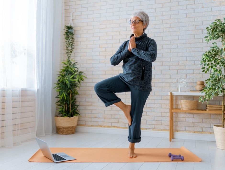 Senior doing tree pose, yoga at home with a laptop and orange yoga mat. 